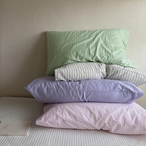 May include: Four pillows in a stack, in shades of green, white, lavender, and pink. The top pillow is light green with black stripes, followed by a striped black and white pillow. The bottom two are solid colours. A book with a red spine is on the bed.