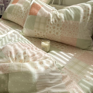 Könnte beinhalten: Ein Patchwork-Bettwäscheset mit einer Mischung aus salbeigrünen, zartrosa und cremefarbenen Quadraten. Die Muster umfassen Blumendrucke, feine Streifen, kleine Schleifen und Karos. Eine Glaskerze mit dem Text Good Night steht auf dem Bett.