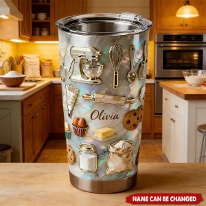 May include: Stainless steel tumbler with a pearlescent finish, adorned with gold-colored baking tools and food illustrations. Features a stand mixer, whisk, rolling pin, cookies, and the name "Olivia". The bottom of the tumbler is silver.