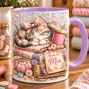 May include: A ceramic mug with a light purple handle and interior, featuring a graphic of a sleeping kitten on a stack of floral fabric, a pink thread spool, a pincushion, a gold thimble, scissors, and a tag with the text Made for Mia.