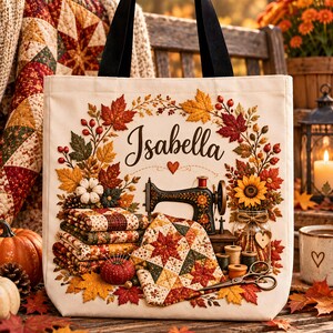 Op de afbeelding: Een crèmekleurige canvas tas met zwarte hengsels, voorzien van een print met een vintage naaimachine, gestapelde lapjes stof, een zonnebloem in een pot en herfstbladeren. De naam Isabella staat in zwarte schrijfletters boven de afbeelding.