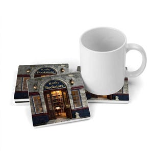 May include: A white ceramic mug and two square coasters featuring a printed image of a stone bookstore storefront with the text Kathy Bookstore above the open wooden doors, revealing a warm interior with bookshelves and a rolling ladder.