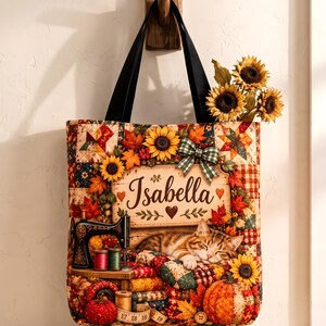 May include: A quilted tote bag featuring a patchwork autumn design with sunflowers, orange maple leaves, a sleeping tabby cat, a vintage sewing machine, and a pumpkin. A central rectangular sign displays the name Isabella in brown script.