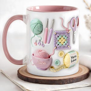 May include: A white ceramic mug with a pink interior and handle, featuring a printed design of mint green yarn, pink and purple crochet hooks, a lavender granny square, scissors, buttons, a pink bowl labeled Lucia, and yellow yarn labeled Yarn Love.