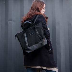 May include: A black tote bag with a leather bottom and the word "ZORKO" printed on the front. The bag has two handles and a zipper closure. The bag is being carried over the shoulder.