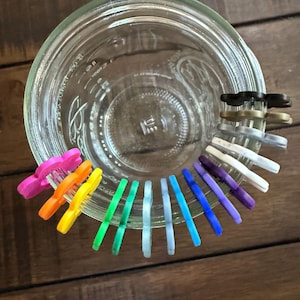 May include: A clear glass jar with a wide opening, surrounded by a variety of colorful, small, plastic clips. The clips are in shades of pink, orange, yellow, green, blue, purple, white, and black. The jar sits on a wooden surface.