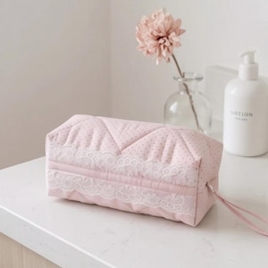 May include: A rectangular, light pink quilted cosmetic bag featuring a subtle dark polka dot pattern and a decorative white lace trim along the front panel. The bag includes a matching pink fabric zipper pull attached to the side closure.