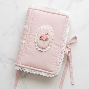 May include: A light pink fabric book cover featuring white lace trim along the edges and a central oval patch with a polka dot pattern. The patch is decorated with three small pink fabric roses and green leaves, and the cover includes pink ribbon ties.