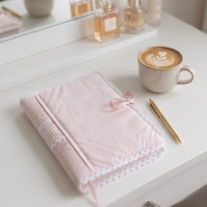 May include: A light pink fabric notebook cover featuring a subtle white pinstripe pattern, white lace trim along the edges, and a small pink ribbon bow closure. A gold-toned pen rests beside the notebook, which is placed on a white surface.