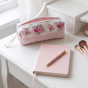 Op de afbeelding: Een rechthoekig gewatteerd etui met een bloemenpatroon van roze rozen en groene bladeren, geaccentueerd met witte kanten randen. Het etui heeft een witte ritssluiting met een roze lipje, vergezeld door een roze notitieboek en een bijpassende pen.