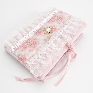 May include: A rectangular fabric case in light pink, featuring a central panel with a pink rose floral pattern. The case is decorated with white lace ruffles along the edges and a central gold-toned oval brooch with a faux pearl. It ties with pink ribbons.