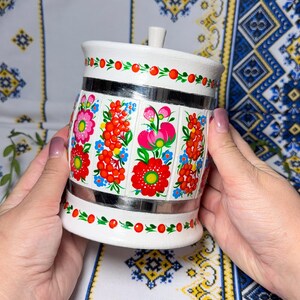 Puede incluir: Tarro de cerámica blanco con tapa, decorado con coloridos motivos florales en rojo, rosa y azul. El tarro tiene bandas plateadas y un pequeño asa en la tapa. El diseño recuerda al arte popular.