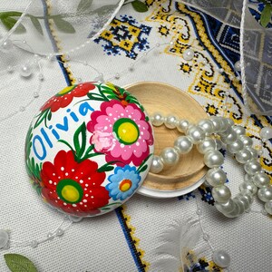 Pode incluir: Um ovo decorativo pintado à mão com o nome "Olivia" em azul, rodeado por flores vermelhas, rosas e azuis com folhas verdes. Uma tigela de madeira e um colar de pérolas brancas estão ao lado do ovo, sobre um tecido estampado.