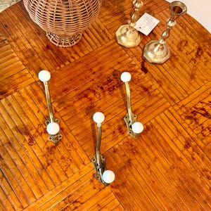 May include: Three brass wall hooks, each featuring two white ceramic knobs at the top and bottom. The hooks have an ornate, curved design with a metallic finish. These functional hardware pieces are arranged on a surface alongside other decorative items.