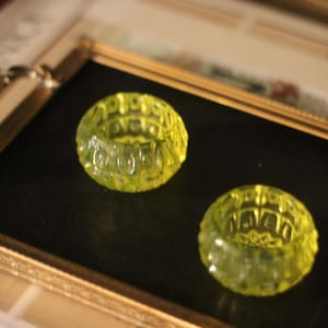 Könnte beinhalten: Zwei kleine, runde, gelb-grüne Glasobjekte mit einem strukturierten, geometrischen Muster. Sie liegen auf einer schwarzen Oberfläche in einem goldgerahmten Display. Die Objekte scheinen dekorativ oder funktional zu sein, möglicherweise zum Halten kleiner Gegenstände.