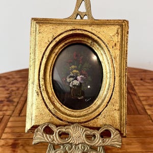 May include: A small rectangular gold-toned frame with an oval center opening containing a dark painting of a floral bouquet. The frame features an ornate top finial and rests on a decorative metal easel stand. The artist signature Tara is visible.