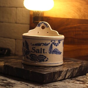 Puede incluir: Un salero de cerámica blanca con diseños florales azules y la palabra "Salt". El salero tiene una parte superior curvada con un agujero para colgar. El fondo incluye una superficie de madera y una lámpara.