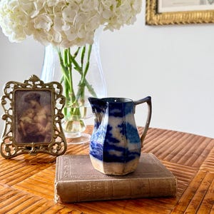 May include: A small, hexagonal ceramic jug featuring a blue and white transferware pattern. The jug has a curved handle and sits atop a vintage hardcover book. The blue design consists of abstract, cloud-like shapes against a white base.