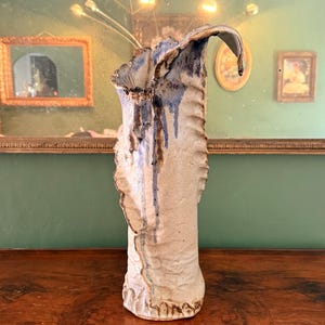 May include: A tall, textured ceramic vase with a cream-colored body and blue accents. The vase has an organic, sculptural shape with a flared top and is displayed on a wooden surface.