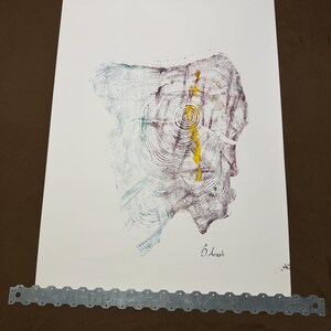 May include: Abstract art print featuring a tree ring design with shades of purple, teal, and yellow on white paper. The artwork is secured with two metal strips at the top and bottom. The artist's signature is visible.