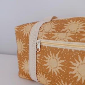 May include: A rectangular fabric bag in a mustard yellow color featuring a repeating pattern of light yellow suns and small dots. The bag includes a cream-colored woven strap and a horizontal zipper closure with a white pull tab on the front.
