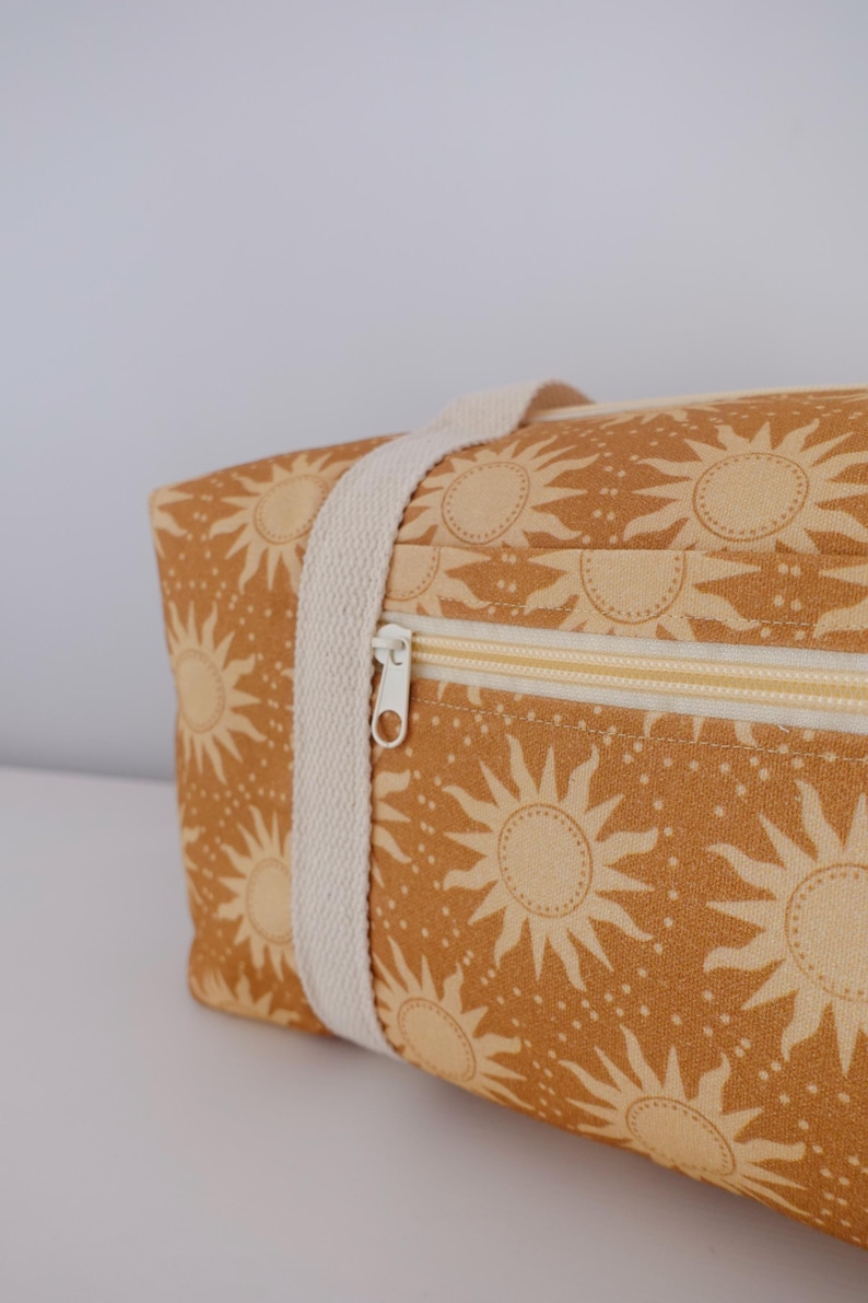 May include: A rectangular fabric bag in a mustard yellow color featuring a repeating pattern of light yellow suns and small dots. The bag includes a cream-colored woven strap and a horizontal zipper closure with a white pull tab on the front.