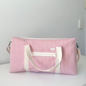 May include: A rectangular duffel bag featuring a light pink and dark red vertical striped pattern. The bag includes two off-white woven fabric handles, a front zippered pocket with an off-white zipper, and a small white label with black text.
