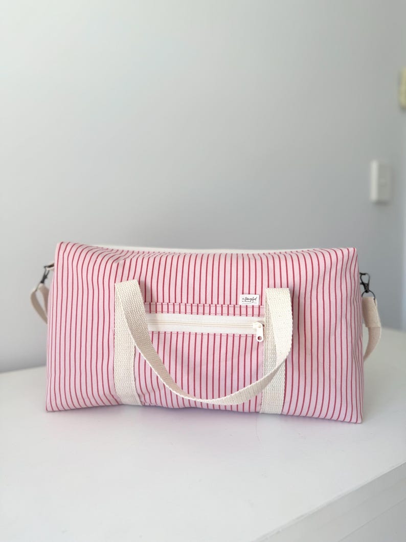 May include: A rectangular duffel bag featuring a light pink and dark red vertical striped pattern. The bag includes two off-white woven fabric handles, a front zippered pocket with an off-white zipper, and a small white label with black text.