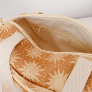 May include: A duffel bag featuring a tan and cream sun pattern with white woven straps. The bag has a top zipper closure and an interior lining with a small zippered pocket. The exterior fabric displays a repeating sun motif with small dots.