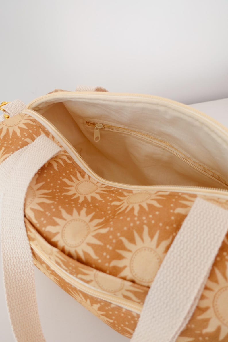 May include: A duffel bag featuring a tan and cream sun pattern with white woven straps. The bag has a top zipper closure and an interior lining with a small zippered pocket. The exterior fabric displays a repeating sun motif with small dots.