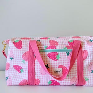 May include: A rectangular duffel bag featuring a pink and white gingham pattern with a repeating strawberry print. The bag includes bright pink fabric handles, a light blue zippered pocket on the front, and a small white label reading n.tangled.