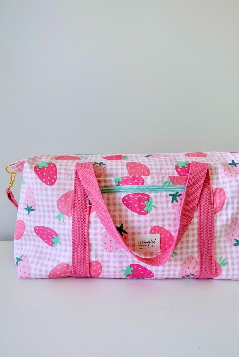 May include: A rectangular duffel bag featuring a pink and white gingham pattern with a repeating strawberry print. The bag includes bright pink fabric handles, a light blue zippered pocket on the front, and a small white label reading n.tangled.
