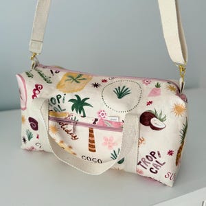 May include: A cream-colored duffel bag featuring a repeating tropical print with pineapples, palm trees, coconuts, and suns. The bag has a pink zippered front pocket, two cream-colored fabric handles, and an adjustable cream-colored shoulder strap.