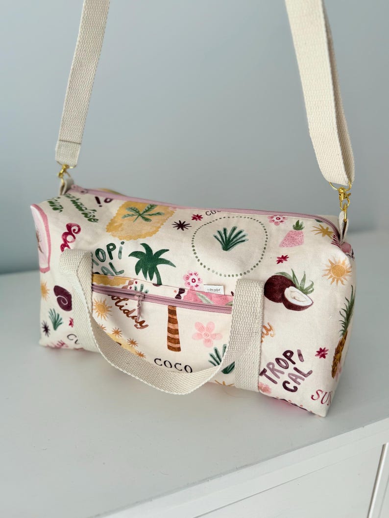 May include: A cream-colored duffel bag featuring a repeating tropical print with pineapples, palm trees, coconuts, and suns. The bag has a pink zippered front pocket, two cream-colored fabric handles, and an adjustable cream-colored shoulder strap.