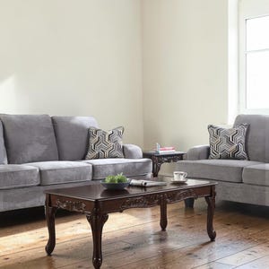 May include: A living room scene featuring a gray sofa, loveseat, and a dark brown coffee table. The coffee table has ornate carvings and curved legs. Decorative pillows with geometric patterns add a touch of style.