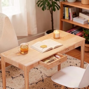 May include: A light-colored wooden desk with a drawer, holding a book, a cup of tea, and a lit candle. The desk is in a well-lit room with a white chair and a patterned rug. A small drawer is open, revealing its contents.