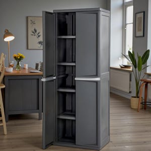 May include: A dark gray storage cabinet with two doors, one open to reveal interior shelves. The cabinet has a rectangular shape and a white handle. The background shows a room with a table, chair, and a window.
