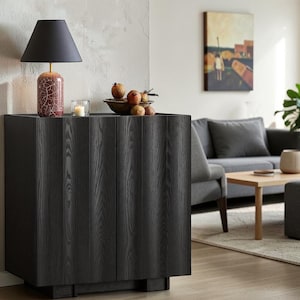 May include: A black wooden cabinet with two doors featuring a vertical grain pattern. On top sits a table lamp with a red crackle-glaze base and a black shade, a small glass candle holder with a white candle, and a bowl filled with brown pomegranates.