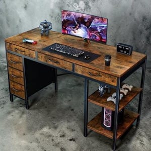 May include: A brown and black gaming desk with a monitor, keyboard, and accessories. The desk has a wood-grain finish, black metal frame, and multiple drawers. A small shelf holds game controllers and a computer tower.