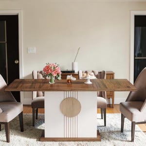 Puede incluir: Una mesa de comedor de madera con base blanca y un círculo decorativo de madera y líneas verticales. La mesa está rodeada de sillas tapizadas y adornada con flores y postres. La habitación tiene una paleta de colores neutros.