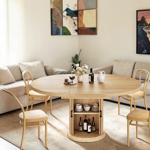 May include: A round light oak dining table with a central storage pedestal featuring two open shelves containing ceramic bowls and four glass wine bottles. Four matching light oak bentwood chairs are positioned around the table.