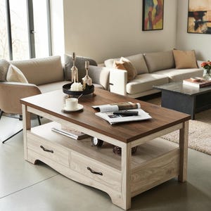 May include: A rectangular coffee table with a dark brown wood-grain top and a light oak-colored base featuring two drawers with black metal handles. The table has an open lower shelf holding books and a small clock, with a magazine on the top surface.