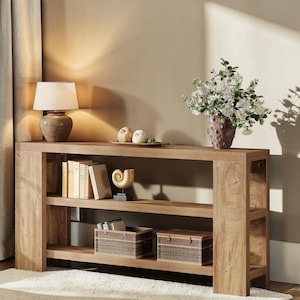 May include: A light brown wooden console table with three shelves. The top shelf holds a lamp, books, and decorative items. The middle shelf has a decorative shell and two woven baskets. A vase of flowers sits on the right.