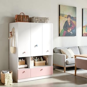 May include: A white storage cabinet with three upper doors featuring small round knobs in pastel colors, two open middle shelves containing baskets and books, and two lower drawers in a soft pink finish. A brown bag hangs on the side.