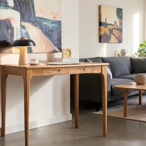May include: A light brown wooden desk featuring four tapered legs and two drawers with integrated semi-circular handles. The desk surface holds a mushroom-shaped lamp with a black shade and light wood base, a small cup, and a square clock.