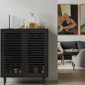 May include: A black wooden cabinet with horizontal slatted doors and four tapered legs. The top surface holds a clear glass decanter filled with amber liquid, a small glass with a stirring rod, and a patterned crystal tumbler glass.