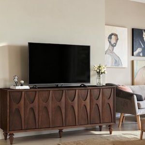 May include: A long, dark brown wooden media console featuring six cabinet doors with a repeating arched cutout pattern. The console stands on six turned legs and supports a large flat-screen television, a small decorative sculpture, and a vase.