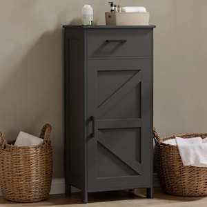 May include: A dark gray bathroom storage cabinet with a drawer and a door featuring a diagonal design. Two woven baskets, one on each side, hold white towels. A small container with toiletries sits on top of the cabinet.