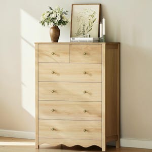 May include: A light-colored wooden dresser with six drawers and brass knobs. The top of the dresser features a vase of flowers, a framed botanical print, candles, and a book. The dresser has a scalloped base and is set against a neutral wall.