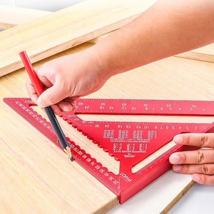 Puede incluir: Una escuadra de velocidad y una regla de aluminio rojas se utilizan para marcar una pieza de madera con un lápiz. La escuadra tiene medidas en pulgadas y tablas de conversión. El lápiz es rojo y negro. La madera es de color marrón claro.
